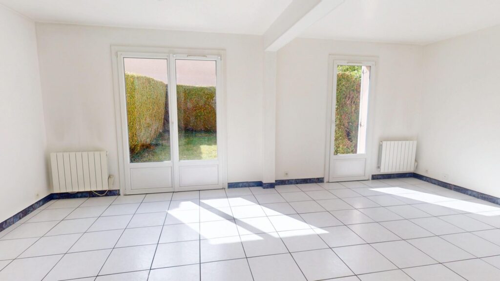 Salon lumineux vide, carrelage blanc et baies vitrées ouvrant sur un jardin.