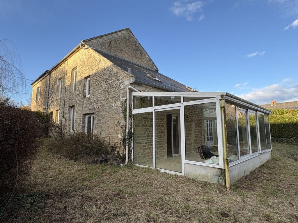 Maison en pierre avec véranda vitrée attenante, vue sur le jardin et façade arrière sous ciel bleu