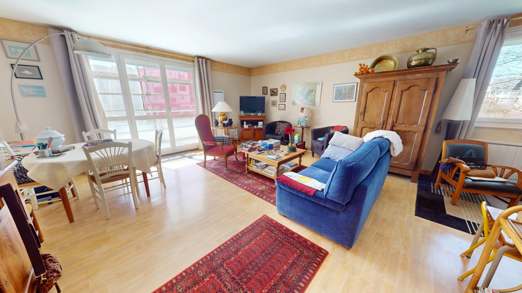 Salon lumineux avec canapé bleu, tapis orientaux, armoire en bois et grande baie vitrée