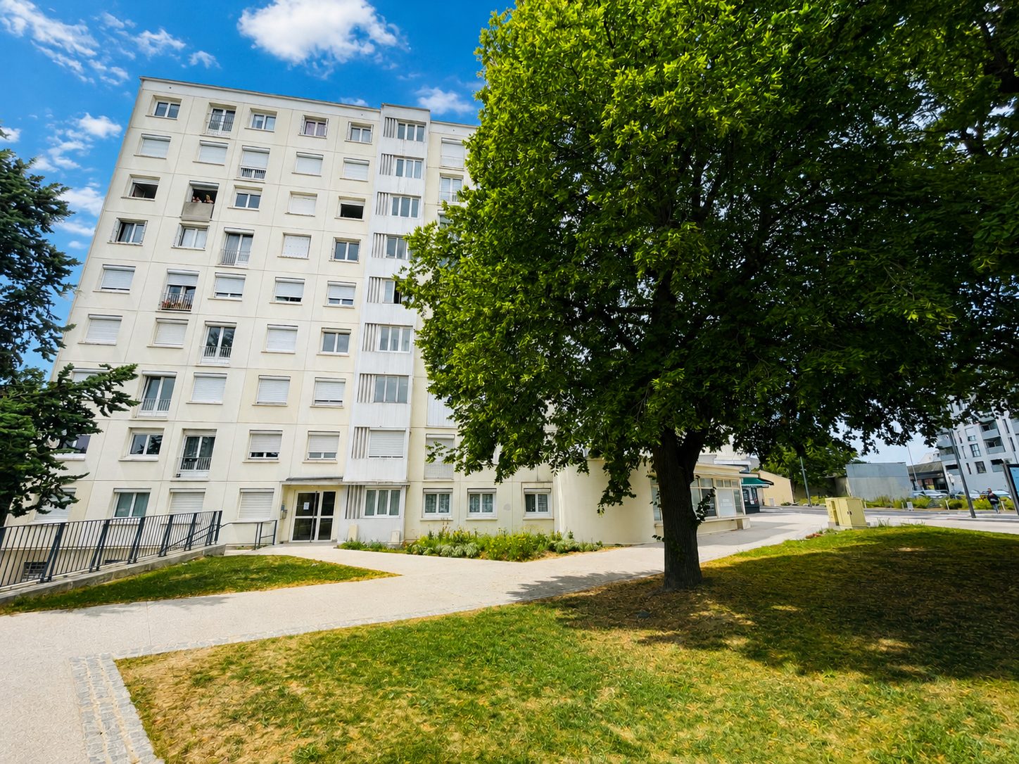 Immeuble résidentiel à façade blanche avec grand arbre ombragé au premier plan, ciel bleu et allée piétonne.