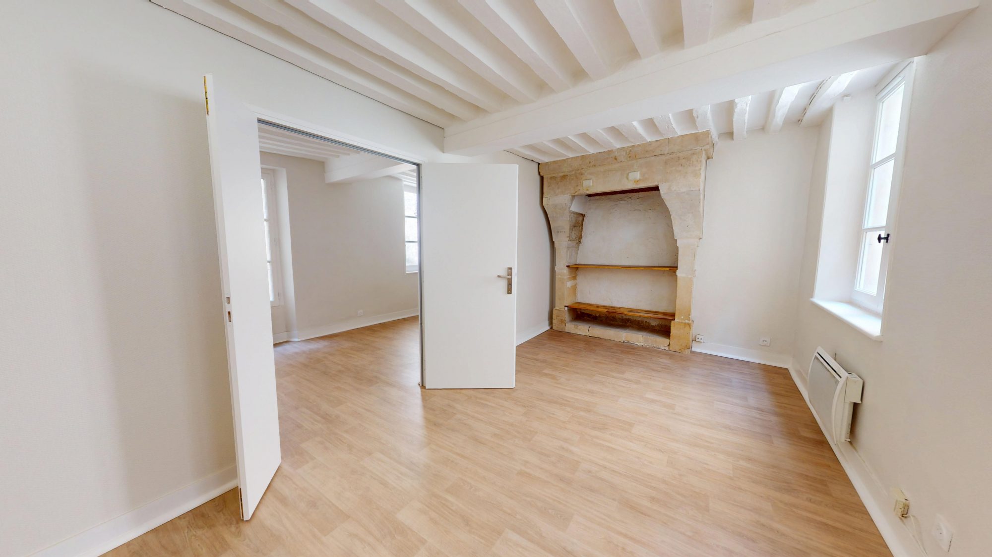 Pièce lumineuse avec poutres apparentes, parquet clair et niche en pierre, porte blanche ouverte